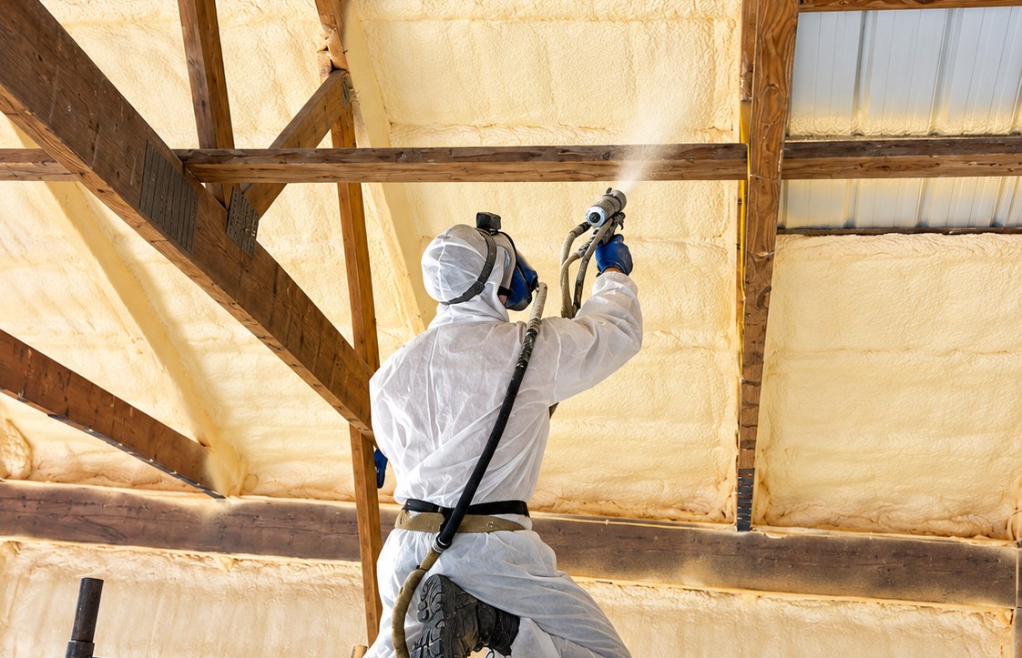 Technician in protective gear spraying foam insulation across a barn ceiling